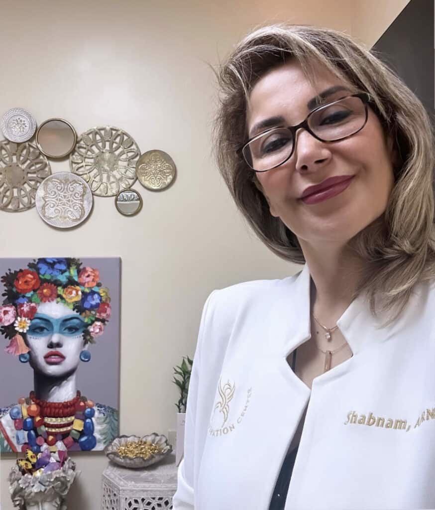 Portrait of Shabnam at Homa Rejuvenation smiling in a treatment room wearing a white coat with decorative wall art in the background.